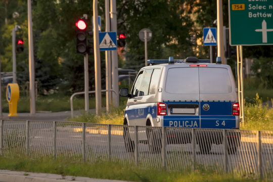 Ostrowiec Świętokrzyski, 23 czerwca 2026, 19:43. Policyjny furgon Volkswagen na skrzyżowaniu z czerwonym światłem. Furgonetka w niebiesko-białym malowaniu stoi na skrzyżowaniu ze znakami drogowymi 