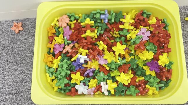 Colorful plastic flower-shaped toy pieces in a yellow tray on a carpet, showing a messy pile of small educational building parts for play and learning. lego