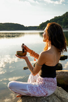 Woman practicing yoga by a calm lake with singing bowl