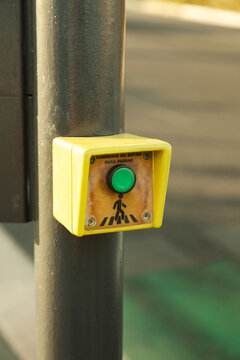 Close-up of a pedestrian crossing push button mounted on a traffic light pole in an urban environment. The accessible crossing control features a green push button with a pedestrian symbol