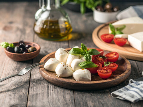A plate of fresh mozzarella balls, cherry tomatoes, and basil sits on a wooden table, with olive oil, olives, and cheese in the background.