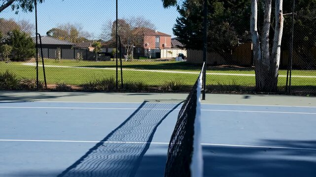 Mesh net of an empty blue tennis court in a quiet suburban Australian park with hard court surface, framing a tranquil backdrop of green parkland, a walking path, and residential homes