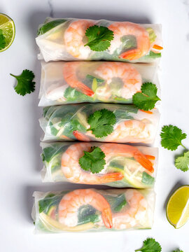 A top-down view of five fresh spring rolls filled with shrimp and vegetables, arranged vertically on a white marble surface. Garnishes of cilantro and lime wedges surround the rolls.