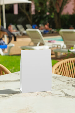 Blank vertical table tent mockup placed on a marble table beside an outdoor swimming pool at a leisure resort. The empty display provides clean copy space for restaurant menus, promotional offers