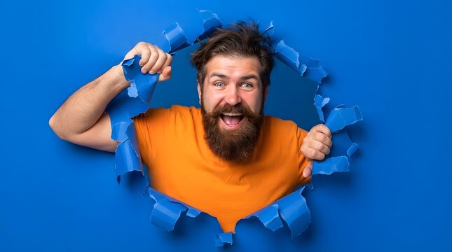 Excited bearded man in orange shirt breaking through a blue paper background looking at the camera