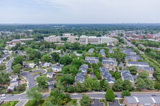 Aerial view of a lush green suburban neighborhood blending into a commercial district in Fairfax, Virginia. Features homes, townhouses, large office buildings, and a distant city skyline.