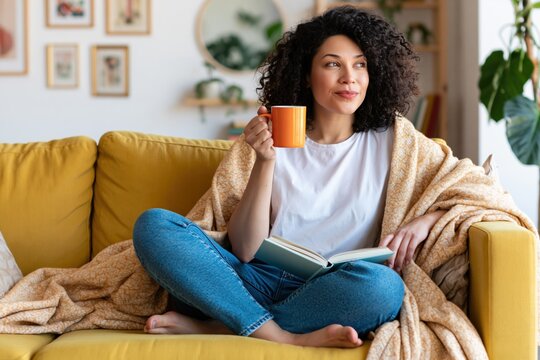 Woman relaxing on a yellow sofa with a cozy blanket reading a book and holding mug
