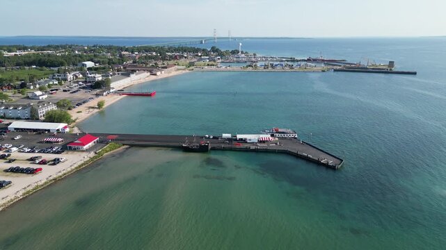 Aerial footage over the historic Mackinac City shoreline. Piers and docks jut from the sandy shoreline for the tour boats to moor. City and bridge alive with adventure and experience