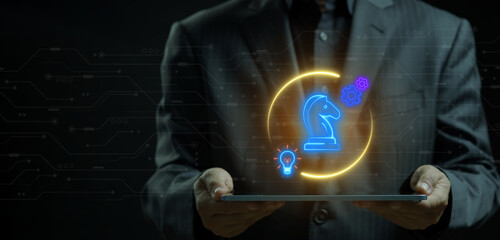 Hands holding tablet displaying glowing chess knight icon inside circle surrounded by lightbulb and gear icons. Cyberpunk neon lighting, strategic business tactics and automated innovation concept. © kobps2