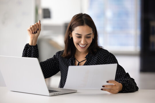 Happy Middle-Eastern business woman reading great news on paper document, celebrate professional success with clenched fist seated at her desk with open laptop. Triumph, career, promotion, achievement