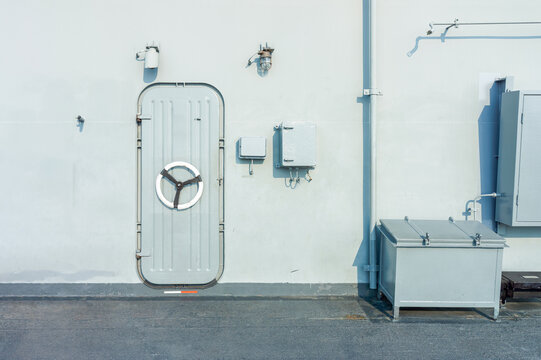 Watertight Hatch Door and Bulkhead Wall on Military Ship Deck. Heavy steel watertight hatch with wheel handle mounted on bulkhead wall with electrical boxes and storage container on naval warship deck