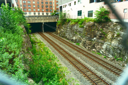 An industrial railway scene showing dual tracks leading into a deep dark tunnel set amidst rocky walls, lush green vegetation, and an urban building exterior.
