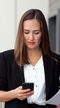Professional businesswoman in a formal suit concentrating on her work, holding papers and using her smartphone for communication and checking information while standing outdoors