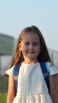 Adorable little girl with a school backpack standing outdoors in the warm sunlight, looking at the camera with a happy and confident smile, ready for her first day of school
