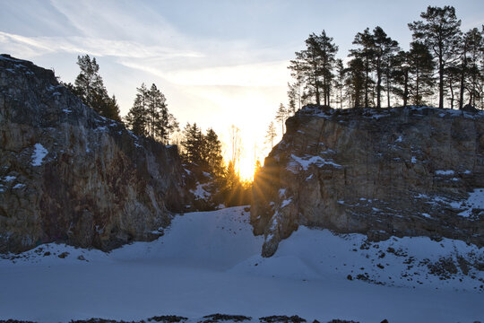 Impressive rock quarry in Sweden during winter with the rising sun peeking through cliffs and pine trees, casting long shadows across the snow-covered ground at Lake arr area.