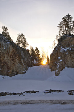 Impressive rock quarry in Sweden during winter with the rising sun peeking through cliffs and pine trees, casting long shadows across the snow-covered ground at Lake arr area.