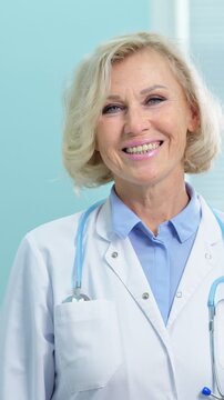 Close up portrait of a friendly senior female doctor in a white lab coat with a stethoscope around her neck, smiling confidently and looking directly at the camera in a clinic setting