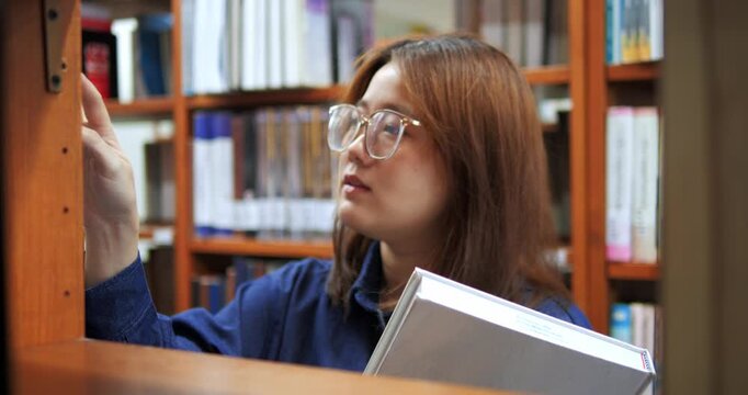 Young woman studies in a library. She wears glasses and holds a book. Her expression shows focus and concentration.