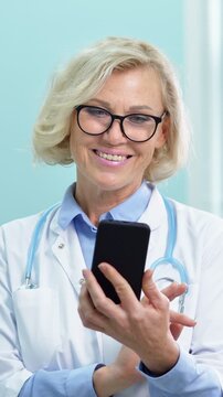 Senior smiling female physician in a white lab coat with a stethoscope holding and using a modern smartphone, scrolling through a medical app for patient information or telehealth communication