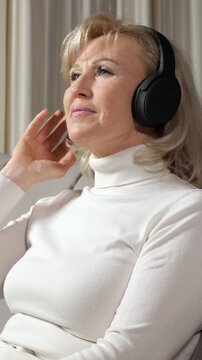 Peaceful senior woman with blonde hair relaxing on a couch at home, wearing wireless headphones and enjoying her favorite music with her eyes closed, feeling calm and serene