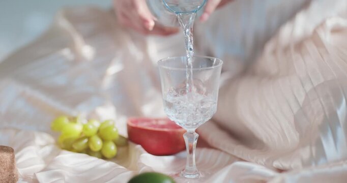 Glass goblet receives fresh water poured from a carafe held by a person in a brightly lit room featuring fresh grapes and a halved grapefruit arranged artistically on a soft textured fabric surface.