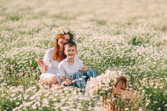 Redhead mother and young son enjoying picnic in sunny chamomile field during bright spring day