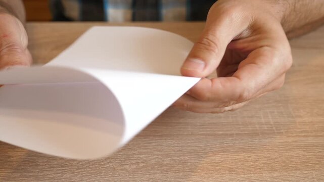 Person folding a white sheet of paper in half by hand on a wooden table in close up. Continuous action with original sound.