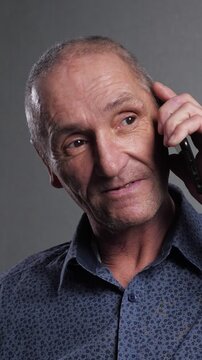 Elderly caucasian man with short gray hair engaged in a phone conversation, his facial expressions shifting from serious to smiling as he listens and responds against a plain gray studio background