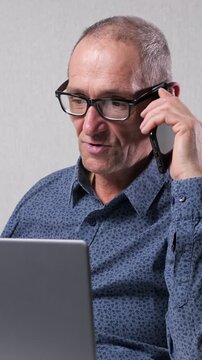 Elderly caucasian man with glasses multitasking in his office, talking on the phone while simultaneously working on his laptop, showcasing modern business communication and productivity