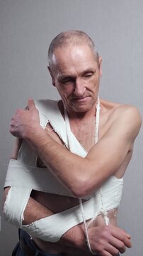 Shirtless senior man with a fractured arm and dislocated shoulder in a white plaster cast is feeling pain and discomfort, illustrating the challenging recovery process after a serious injury
