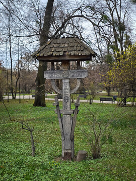 Traditional carved wooden wayside cross (troiță) with shingle roof standing in a green park surrounded by trees.