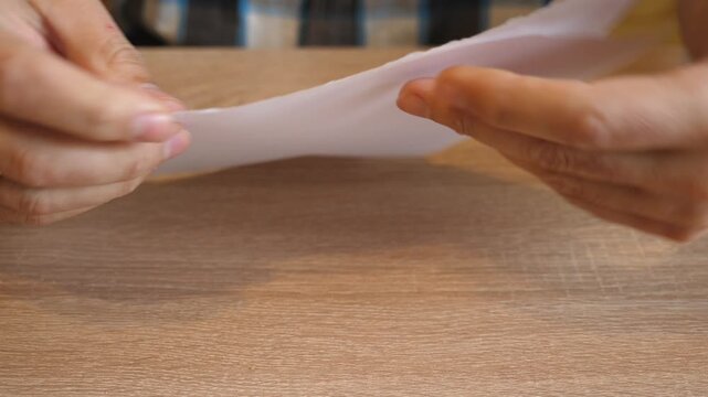 Person tearing a white sheet of paper into pieces by hand above a wooden table in close up. Continuous action with original sound.