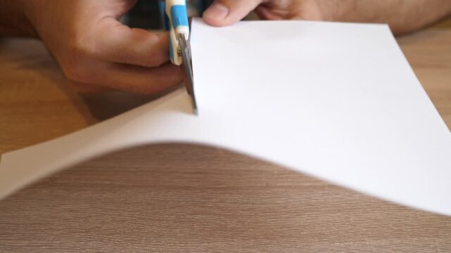 Person cutting a white sheet of paper with scissors on a wooden table in close up. Continuous action with original sound.