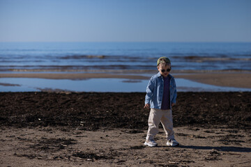 Young boy wearing sunglasses and denim jacket stands on sandy beach with calm sea and clear sky in the background, enjoying a sunny day © Riga Frames