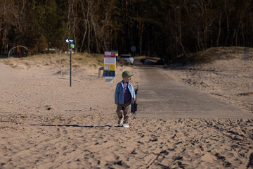 Young child in a blue jacket and cap stands on sandy beach path with signs and trees in the background at Jurmala, Latvia © Riga Frames