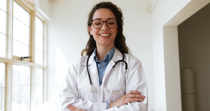 Young medical professional woman in glasses, lab coat and stethoscope posing in hospital room, medical office, looking at camera with hands folded, getting happy, smiling for head shot