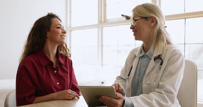 Friendly blonde female doctor in glasses showing screening image on tablet to positive young patient woman, explaining electronic medical report, shaking hands after consultation