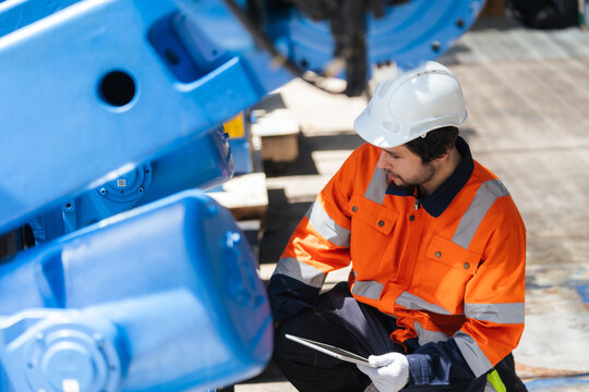 Professional engineer kneels near refurbished industrial robot arm, checking machinery condition with tablet for robotics system integration, equipment maintenance, plant engineering, installation.