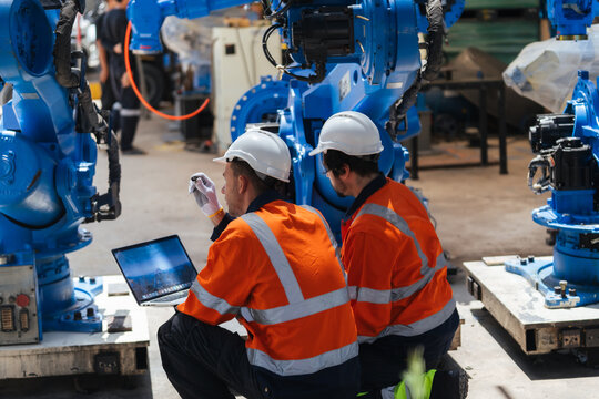 Team technicians review blue industrial robots in factory using laptop, checking machinery condition for equipment maintenance, installation planning, engineering collaboration, automation.