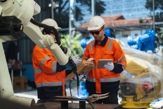 Selective focus, Welding robot torch operate welding sparks on clamped steel fixture test while two robotic technicians using teach pendant and digital tablet for calibration, robotic programming.