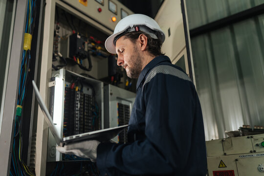 Electrical engineer using laptop near open electrical cabinet, conducting control system diagnostics, facility maintenance management, workplace safety compliance. industrial.