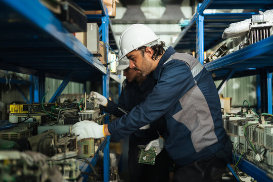 Maintenance technicians inspect robotic arm spare parts on warehouse shelves, checking circuit boards and industrial equipment for parts identification, repair planning, inventory control.