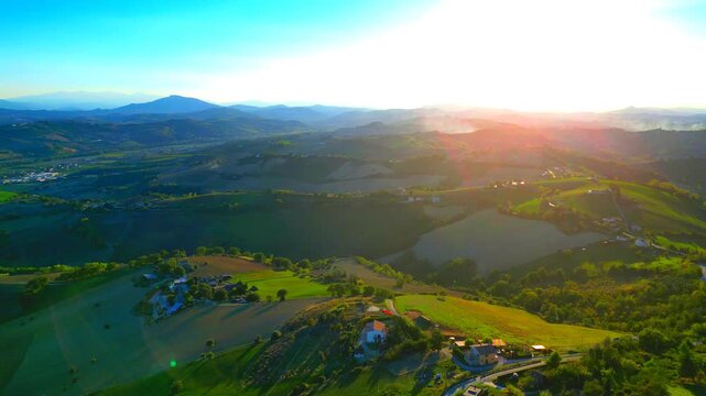Gracious aerial pullback pan left shot from Monterubbiano, afternoon sunlight imbuing the gentle Marche hillscapes with spiritual quality, among smokes, trees, buildings, sunflares, distant mountains