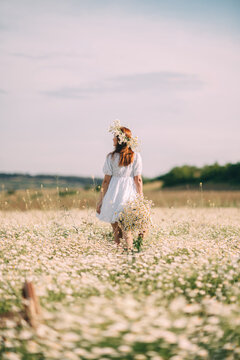 Redhead girl walking through spring meadow wearing Matricaria chamomilla crown and holding a large bouquet of wildflowers