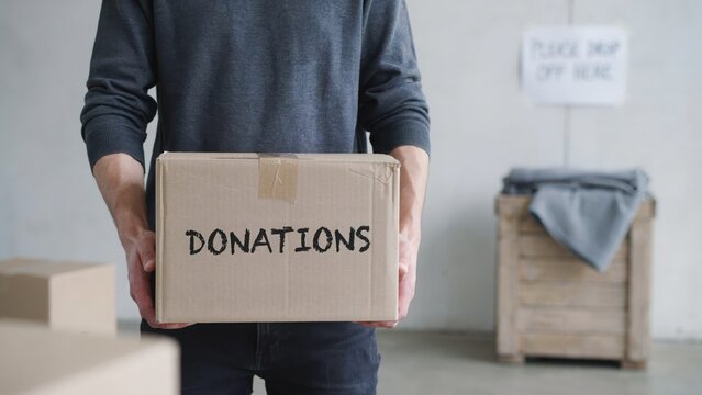 Person holding cardboard box labelled donations standing indoors ready to give items for charity work aid relief support and community help social programs.