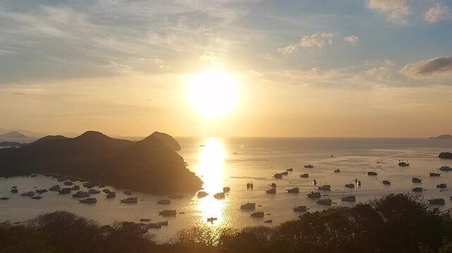 Pemandangan menakjubkan dari ketinggian yang memperlihatkan matahari terbenam keemasan nan berkilau di atas pelabuhan laut tropis. Matahari yang cerah memantulkan cahaya yang hidup di permukaan air, m