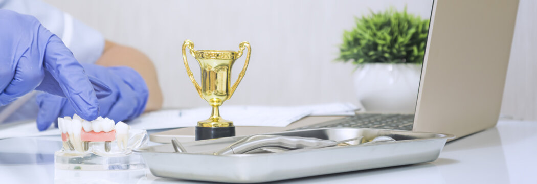 Dentist hand in blue glove pointing at anatomical jaw model with implants. Award winning dental clinic concept with golden trophy cup on desk.