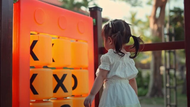 Curious little Asian toddler girl playing with large Tic-Tac-Toe game blocks at an outdoor playground during golden hour. Concentrated child learning through play in a park at sunset.