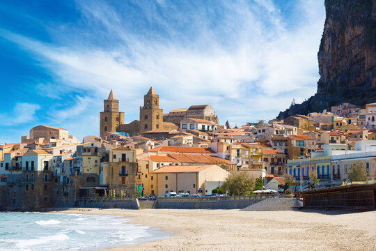 Panoramic view of Cefalu in Sicily, Italy, with Norman Cathedral of Cefalu, historic old town buildings, sandy beach and Tyrrhenian Sea shoreline beneath dramatic clouds and towering La Rocca cliff