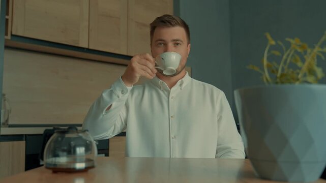 A man starts his serious day with a cup of coffee. A handsome man in a white shirt drinks his favorite coffee from a cup at home and is ready to conquer the world.